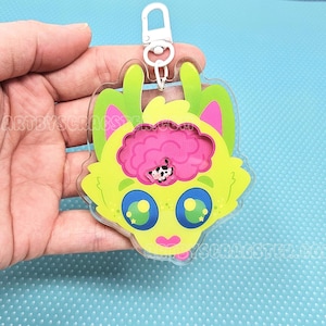 May include: A clear acrylic keychain featuring a cartoon cat with a brain design. The cat has a lime green face, pink ears, and blue eyes. The brain is pink with a black and white pattern. The keychain has a silver clasp.