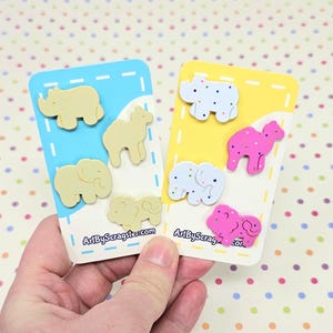 Puede incluir: Dos juegos de pines con forma de galletas de animales. El primer juego es beige y presenta un rinoceronte, un camello y dos elefantes. El segundo juego es rosa y blanco con un rinoceronte, un camello y dos elefanten. Los pines están en una tarjeta con el texto "ArtByScragster.com".