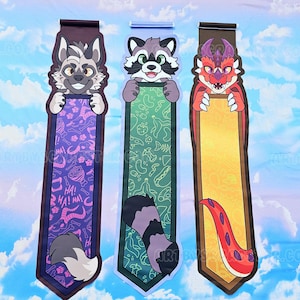 May include: Three illustrated bookmarks with cartoon animal characters. The first features a wolf, the second a raccoon, and the third a dragon. Each bookmark has a unique design with a tail at the bottom and a matching character at the top.