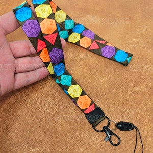 Puede incluir: Un cordón negro con un patrón de dados de colores. El cordón tiene un clip negro y un cierre de seguridad negro. Los dados son de colores arcoíris y tienen números en ellos.
