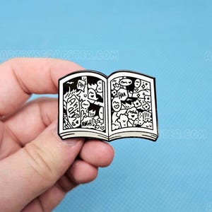 May include: A black and white enamel pin in the shape of an open book. The pages are filled with doodle art, including faces, speech bubbles, and the words "Ah!" and "Raugh!". The pin has a glossy finish and a black border. The website address "ARTBYSCAGSTER.COM" is visible.