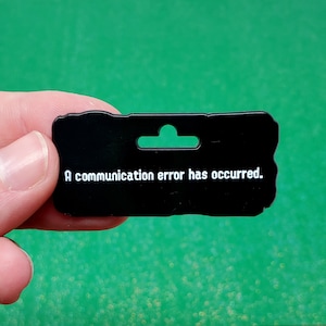 May include: A black rectangular tag with a green hanging hole. The text "A communication error has occurred" is printed in white. The tag is held by a person's hand against a green, glittery background.