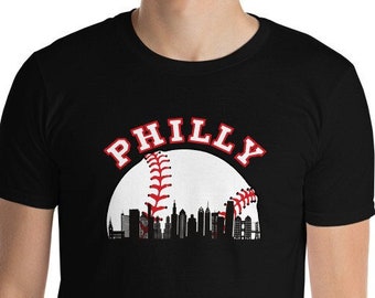 Może przedstawiać: Czarny T-shirt z białym nadrukiem baseballu i tekstem "PHILLY" w kolorze czerwonym. Nadruk baseballu przedstawia sylwetkę panoramy Filadelfii.