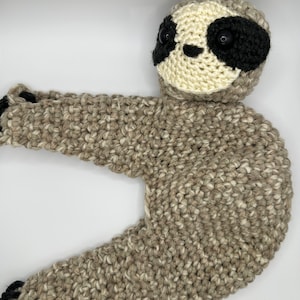 May include: A crocheted sloth toy with brown and white fur, black eyes, and a white face. The sloth is lying on its back with its arms and legs outstretched.