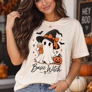May include: A cream-colored short-sleeve t-shirt featuring a graphic of a ghost wearing a black witch hat with an orange bow, holding a coffee cup, and standing next to a pumpkin. The text "Basic Witch" is printed below the ghost illustration.