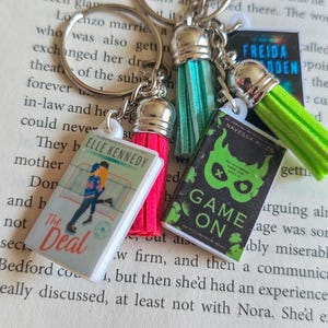 May include: Three metal keychains featuring miniature book covers. One shows The Deal by Elle Kennedy with a pink tassel, one shows Game On by Navessa Allen with a green tassel, and one shows a blue book cover with a teal tassel.