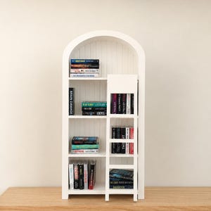 May include: A white arched bookshelf with a vertical sliding ladder, featuring multiple shelves filled with miniature books. The books are arranged in stacks and rows, displaying various titles and colorful spines on the white wooden structure.