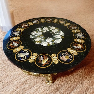 Puede incluir: Una mesa redonda de mármol negro con un borde dorado y una corona de flores blancas en el centro. La mesa está decorada con un borde de conchas marinas y coral.