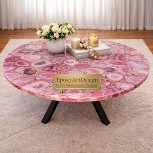 May include: A circular coffee table featuring a pink agate stone surface. The table has a dark base and is adorned with a white vase of flowers, candles, and books. The table has the text "EpoxyArtDesign Luxury Natural Stone Creations".