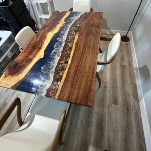 May include: A rectangular wooden dining table with a river-like design. The table features a dark brown wood grain with a blue epoxy resin river, and small stones. The table is surrounded by white chairs with wooden frames.