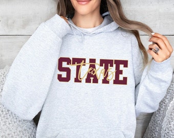 May include: A light gray hoodie with the word "STATE" in maroon block letters and "Texas" in a gold script font. The hoodie has a drawstring hood and a front pocket.