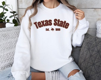 May include: White sweatshirt with the text "Texas State" in maroon arched lettering. Below the text is "Est. 1899" and a maroon bobcat logo. The sweatshirt is a crew neck style.