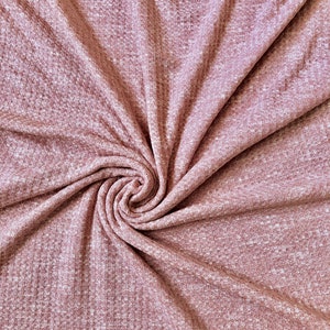 May include: A close-up of a soft, pink waffle knit fabric with a textured surface. The fabric is folded and twisted, creating a swirling pattern.