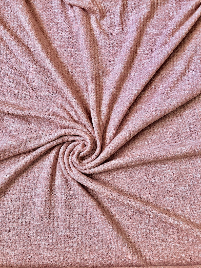 May include: A close-up of a soft, pink waffle knit fabric with a textured surface. The fabric is folded and twisted, creating a swirling pattern.