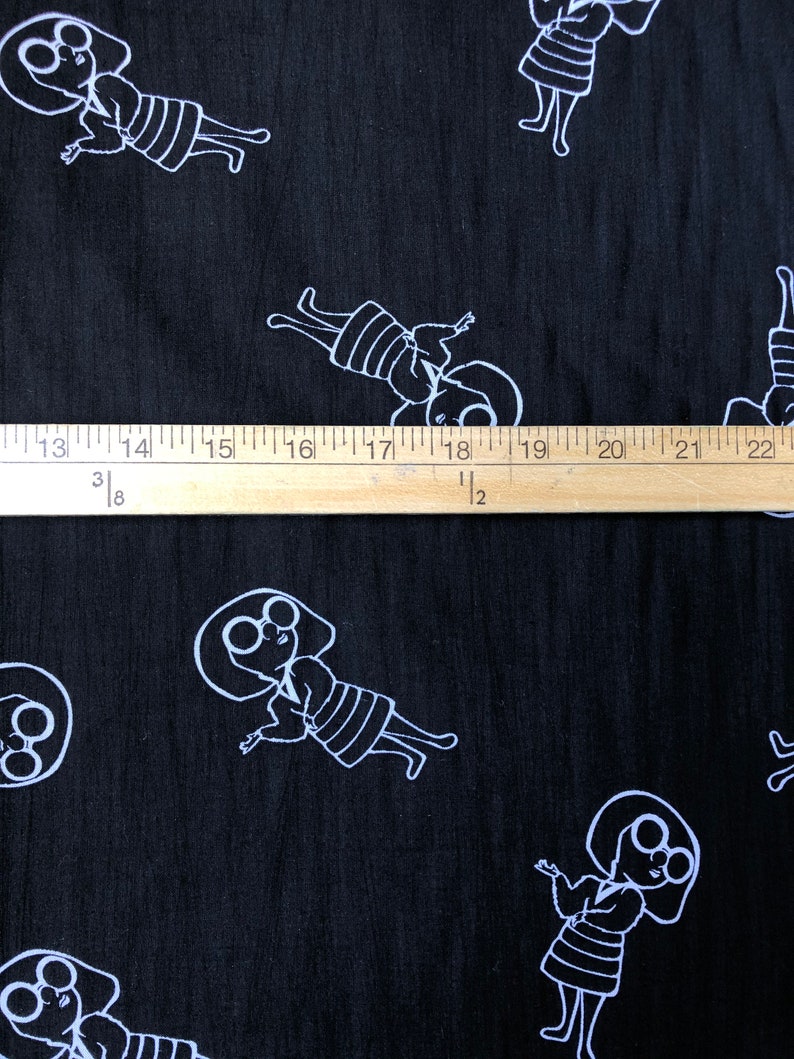 May include: Black fabric with a white repeating pattern of a stylized person with large round glasses. The fabric is measured with a wooden ruler.