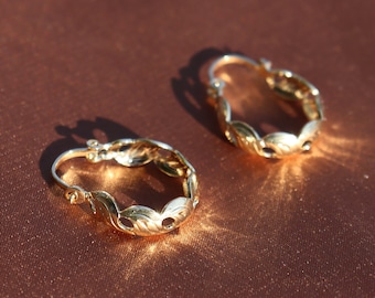 May include: A pair of gold hoop earrings with a delicate, leaf-shaped design. The earrings are on a brown surface.