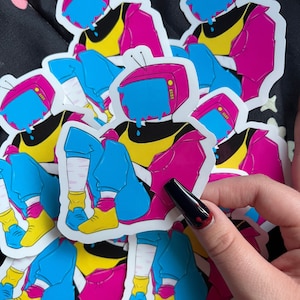 Op de afbeelding: Een stapel kleurrijke stickers met een cartoonfiguur met een televisie als hoofd. Het personage draagt een roze jas, een geel shirt, een blauwe jeans en witte sneakers. De televisie druppelt blauwe verf.