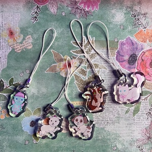 Puede incluir: Una colección de colgantes acrílicos iridiscentes con varios animales y personajes de dibujos animados, incluyendo un hada, una ardilla, un gato y dos cachorros. Cada colgante tiene un cordón blanco para sujetarlo a un teléfono o accesorio.