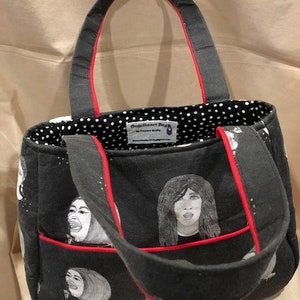 K&ouml;nnte beinhalten: Eine schwarze Tragetasche mit rotem Besatz und einem wei&szlig;en Punktmusterfutter. Die Tasche hat einen schwarz-wei&szlig;en Aufdruck von Gesichtern. Die Tasche hat ein Etikett mit der Aufschrift "Fringe The Art Design".