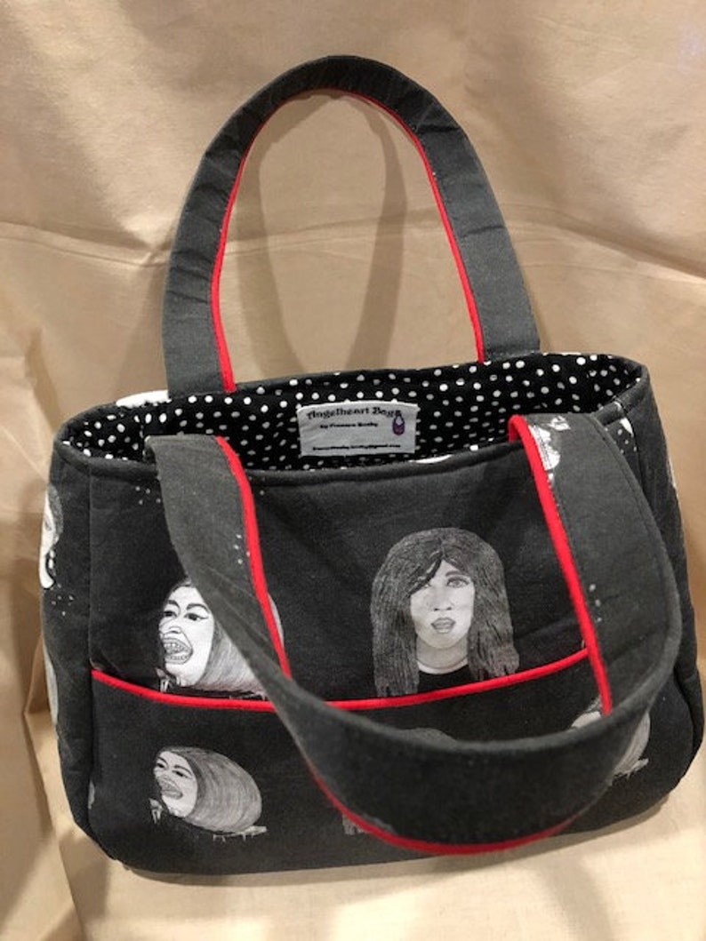 K&ouml;nnte beinhalten: Eine schwarze Tragetasche mit rotem Besatz und einem wei&szlig;en Punktmusterfutter. Die Tasche hat einen schwarz-wei&szlig;en Aufdruck von Gesichtern. Die Tasche hat ein Etikett mit der Aufschrift "Fringe The Art Design".