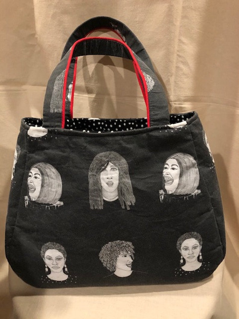 K&ouml;nnte beinhalten: Schwarze Tragetasche mit rotem Besatz an den Griffen. Die Tasche hat ein sich wiederholendes Muster aus schwarz-wei&szlig;en Illustrationen von Frauenportr&auml;ts.