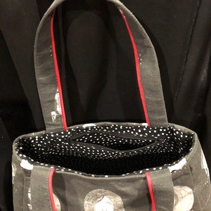 K&ouml;nnte beinhalten: Eine schwarze Canvas-Tragetasche mit rotem Besatz und einem wei&szlig;en, gepunkteten Futter. Die Tasche hat ein sich wiederholendes Muster aus schwarz-wei&szlig;en Gesichtsillustrationen.