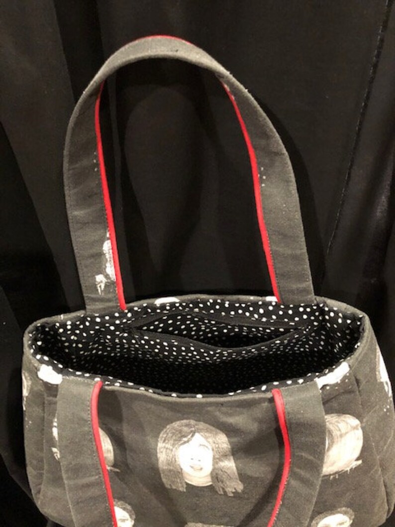 K&ouml;nnte beinhalten: Eine schwarze Canvas-Tragetasche mit rotem Besatz und einem wei&szlig;en, gepunkteten Futter. Die Tasche hat ein sich wiederholendes Muster aus schwarz-wei&szlig;en Gesichtsillustrationen.