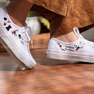May include: White canvas shoes with embroidered floral designs and the date 05/30/2023 and the text "Mrs. Palmez" on one shoe. The other shoe has a black cat and floral embroidery.