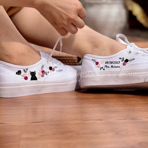 May include: White canvas shoes with floral embroidery and a black cat design on one shoe. The other shoe has the date 03/30/2023 and the text "Mrs. Pel&aacute;ez" embroidered on it.