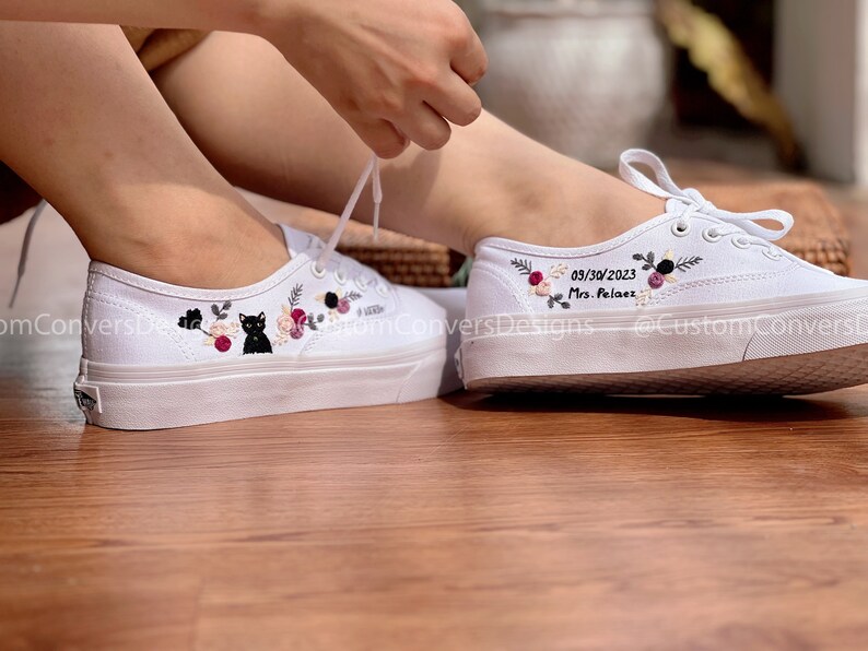 May include: White canvas shoes with floral embroidery and a black cat design on one shoe. The other shoe has the date 03/30/2023 and the text "Mrs. Pel&aacute;ez" embroidered on it.