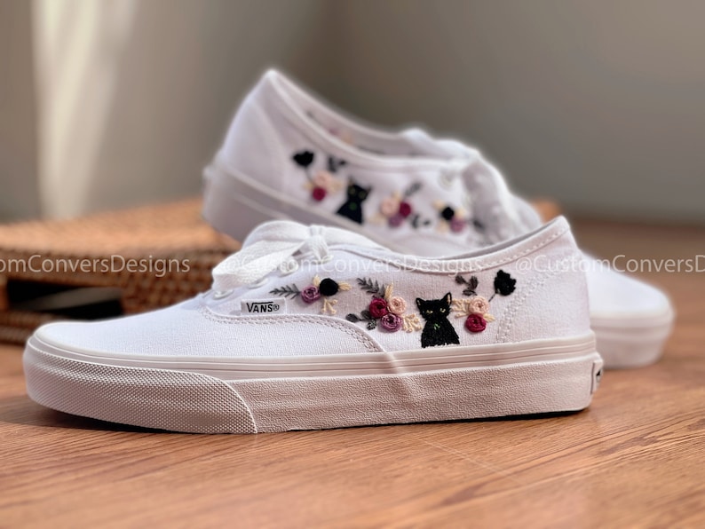 May include: White canvas Vans sneakers with black embroidered cats and flowers. The text "VANS" is visible on the side of the shoe.