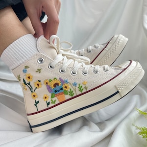 Puede incluir: Zapatillas blancas de lona de caña alta con flores bordadas y un personaje de dibujos animados. Las zapatillas tienen una suela de goma blanca y costuras rojas.