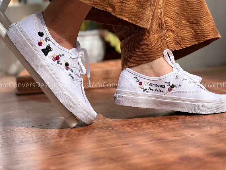 May include: White canvas shoes with embroidered floral designs and the date 05/30/2023 and the text "Mrs. Palmez" on one shoe. The other shoe has a black cat and floral embroidery.