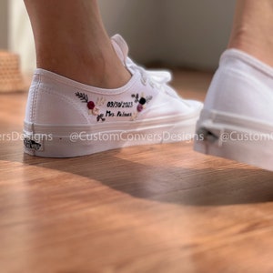 May include: White canvas sneakers with embroidered flowers and the text "09/30/2023 Mrs. Feliciano" on the side. The shoes are worn by a person standing on a wooden floor.