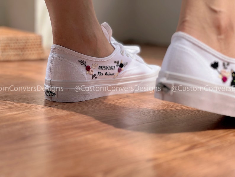 May include: White canvas sneakers with embroidered flowers and the text "09/30/2023 Mrs. Feliciano" on the side. The shoes are worn by a person standing on a wooden floor.