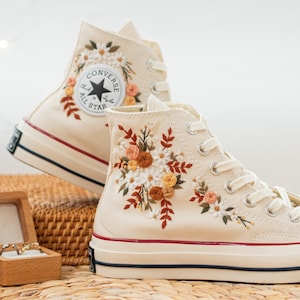May include: Cream-colored high-top sneakers with floral embroidery. The sneakers feature white daisies, orange and peach roses, and green leaves. The Converse logo is visible on the side. The soles have a red stripe.