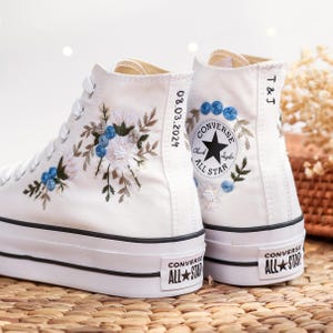 Peut inclure: Une paire de baskets blanches montantes avec des broderies florales bleues. Les baskets ont des finitions noires et un logo noir "Converse All Star" sur le côté. Une basket a la date "08.03.2024" brodée sur le côté, et l'autre a les initiales "T & J" brodées sur le côté.