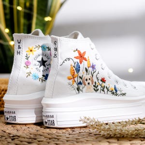 May include: White Converse high-top sneakers with embroidered floral and dog designs. The sneakers feature a black Converse All Star logo and a thick white sole. The embroidery includes colorful flowers and a small dog, adding a personalized touch to the footwear.