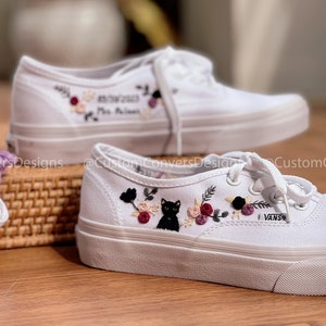 May include: A pair of white canvas shoes with embroidered floral designs and a black cat. The shoes have black laces and the word "VANS" is embroidered on the side. One shoe has the text "07.07.2023" and "Mrs. Williams" embroidered on it.