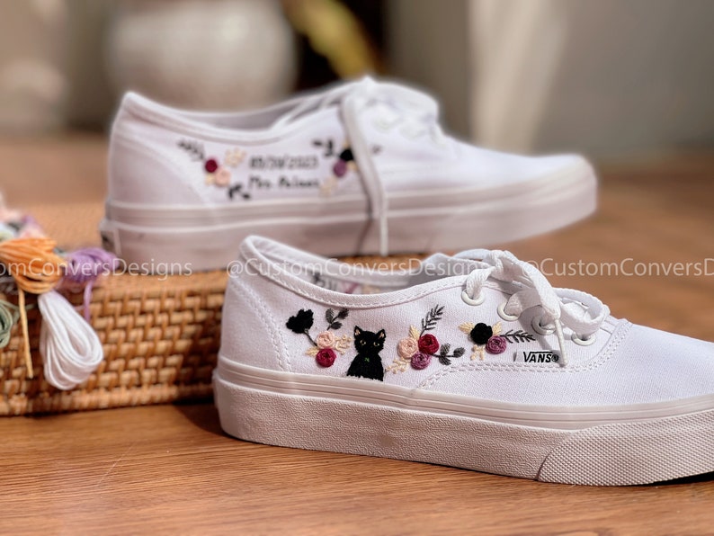 May include: A pair of white canvas shoes with embroidered floral designs and a black cat. The shoes have black laces and the word "VANS" is embroidered on the side. One shoe has the text "07.07.2023" and "Mrs. Williams" embroidered on it.