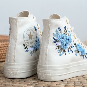 Peut inclure: Des baskets Converse blanches montantes avec des broderies florales bleues. Les baskets ont une semelle en caoutchouc blanche et un œillet en métal doré. Le logo Converse All-Star est visible sur le côté de la chaussure.