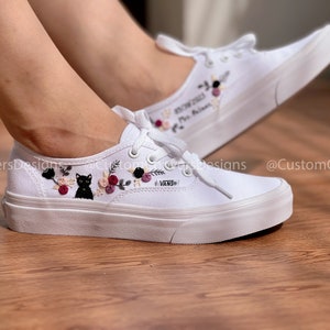 May include: White canvas low-top sneakers with black embroidered cat and floral design. The shoes have a white rubber sole and the word "VANS" is embroidered on the side.