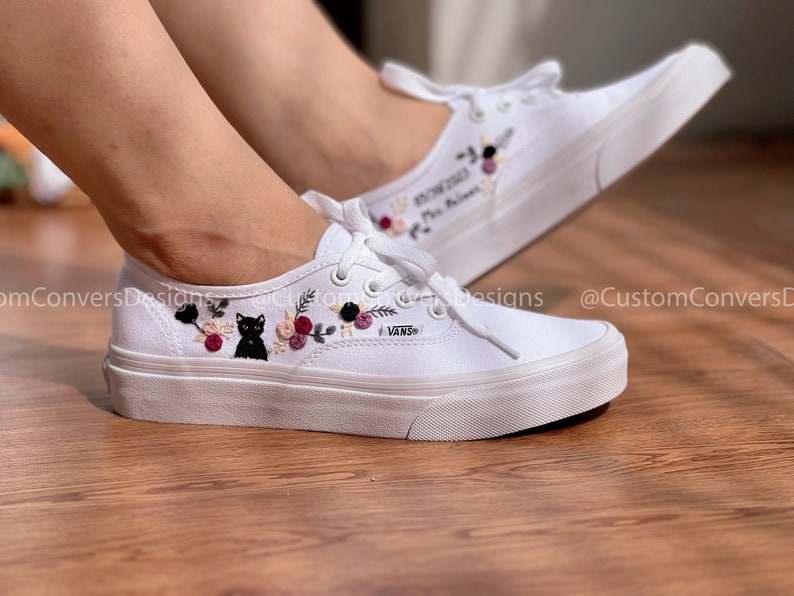 May include: White canvas low-top sneakers with black embroidered cat and floral design. The shoes have a white rubber sole and the word "VANS" is embroidered on the side.