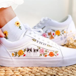 Puede incluir: Zapatillas blancas de lona con flores bordadas, dos gatos y la fecha 2-22-24. Las zapatillas las lleva una persona que lleva calcetines blancos con girasoles bordados.