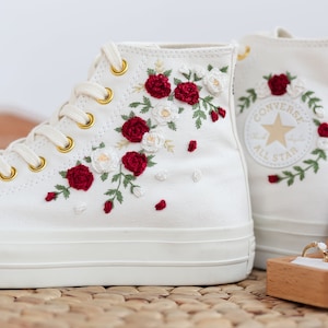 Peut inclure: Des baskets blanches montantes avec des fleurs brodées rouges et blanches. Les baskets ont des œillets dorés et le logo Converse All Star est visible sur le côté.