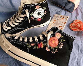 May include: A pair of black high-top Converse sneakers with white laces and embroidered flowers. The sneakers have a white rubber sole and the Converse All Star logo is visible on the side. The sneakers are on a blue surface.