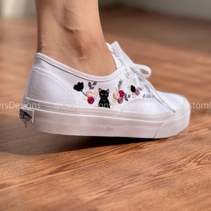 May include: White canvas low-top sneakers with black embroidered flowers and a black cat. The shoes are on a wooden floor.