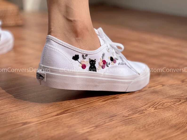 May include: White canvas low-top sneakers with black embroidered flowers and a black cat. The shoes are on a wooden floor.