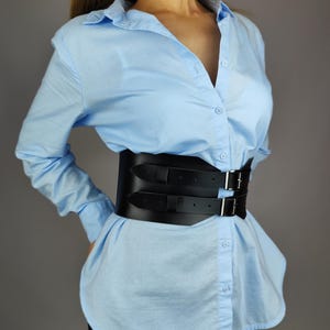Puede incluir: Un cintur&oacute;n cors&eacute; de cuero negro con hebillas dobles y correas. El cintur&oacute;n se lleva sobre una camisa de botones azul claro. El texto "Handmade PIRUS CRAFT STUDIO ETSY" es visible en un c&iacute;rculo blanco.