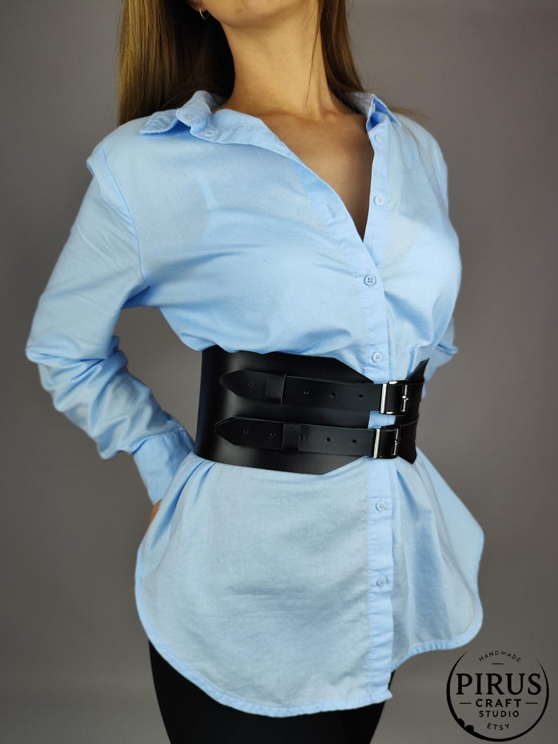 Puede incluir: Un cintur&oacute;n cors&eacute; de cuero negro con hebillas dobles y correas. El cintur&oacute;n se lleva sobre una camisa de botones azul claro. El texto "Handmade PIRUS CRAFT STUDIO ETSY" es visible en un c&iacute;rculo blanco.
